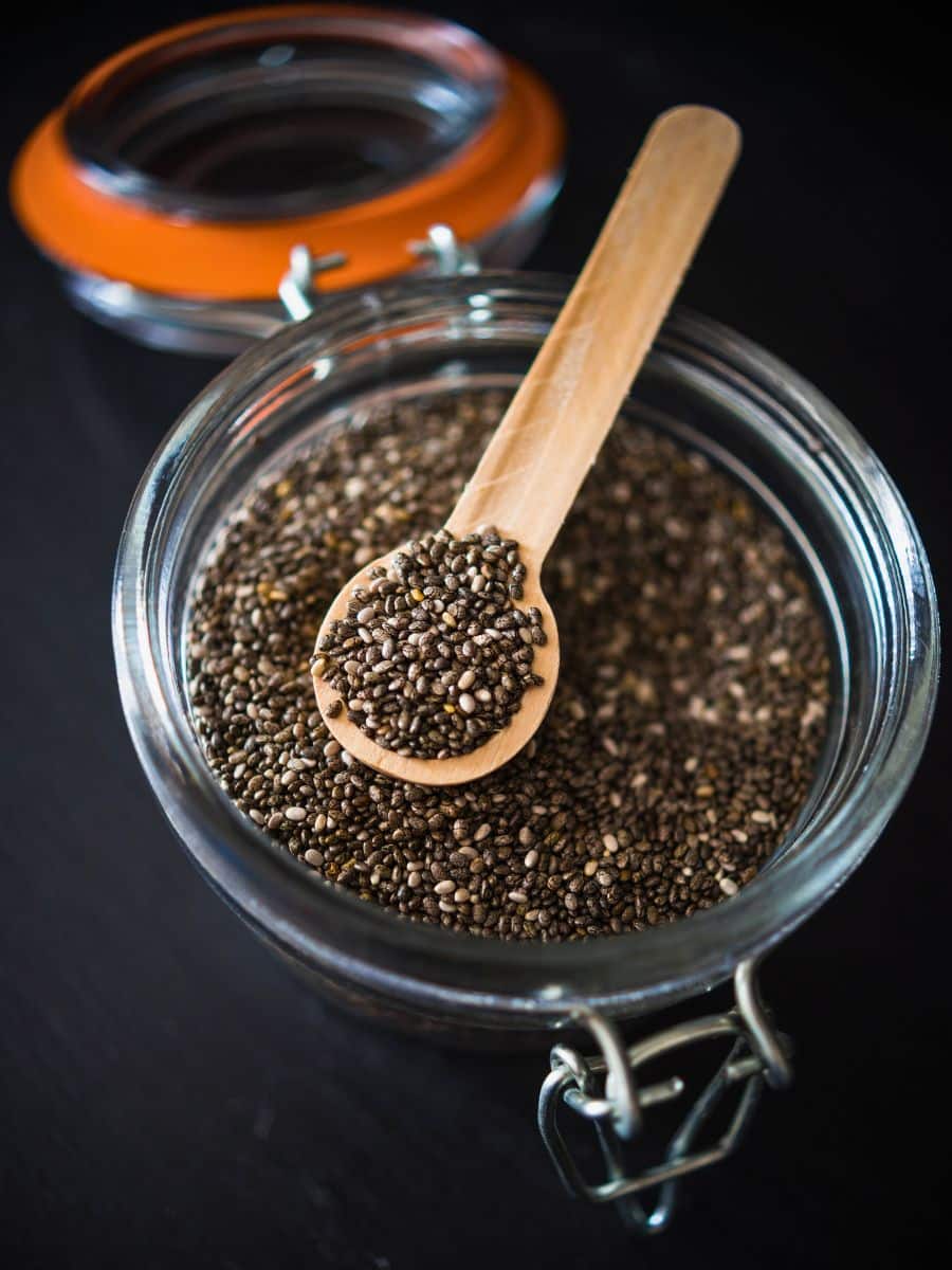 A glass jar filled with chia seeds, with a wooden spoon resting on top, also containing chia seeds. The jar has a metal clasp and an orange seal.