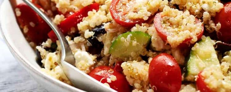 Close-up of a quinoa salad with cherry tomatoes, cucumber, feta cheese, and a spoon in a bowl.