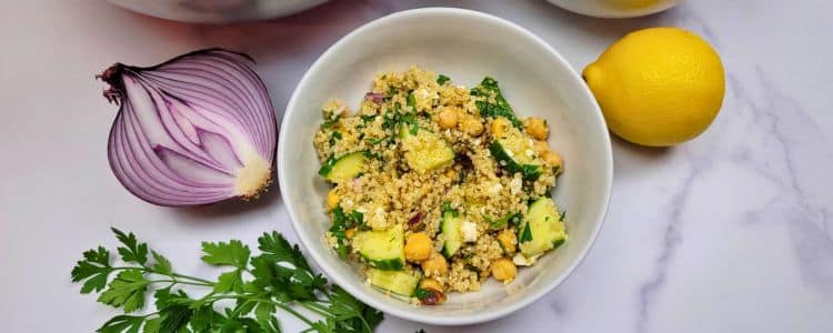 Bowl of quinoa salad with chickpeas, cucumber, and herbs beside half a red onion, a lemon, and parsley on a marble surface.