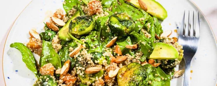 A plate of salad with quinoa, Brussels sprouts, avocado slices, almonds, and spinach leaves, served with a fork on the side.