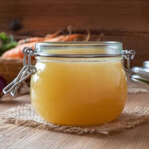 A glass jar filled with homemade chicken stock on a wooden counter with carrots and other vegetables surrounding it.