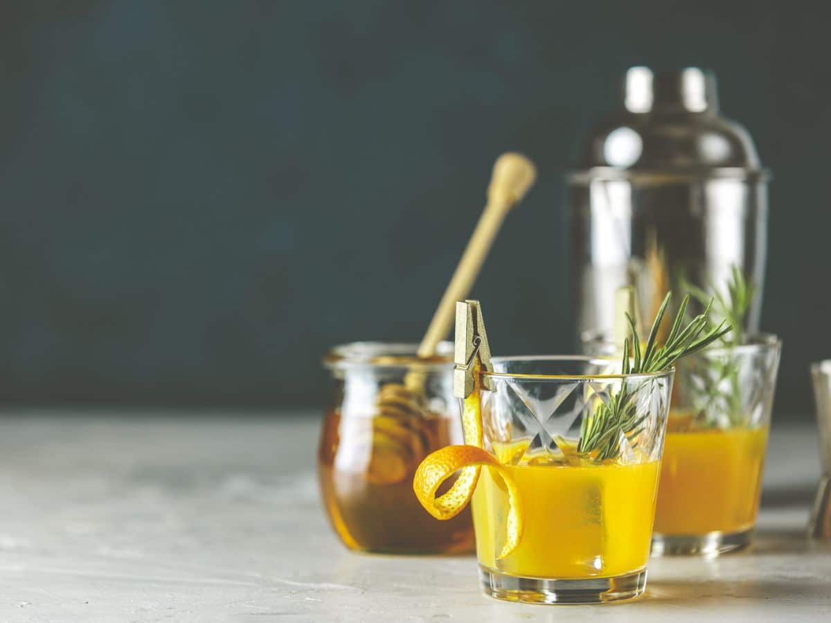 Two orange cocktails garnished with rosemary and orange peel in short glasses, with a cocktail shaker in the background on a textured surface.