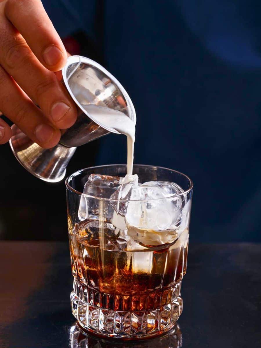 A person pouring cream into a glass of iced coffee, creating a swirling effect. the background is dark, highlighting the drink.