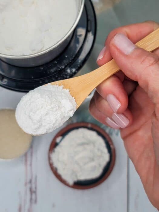 A hand holding a wooden spoon with white powder, likely flour, above a small bowl.