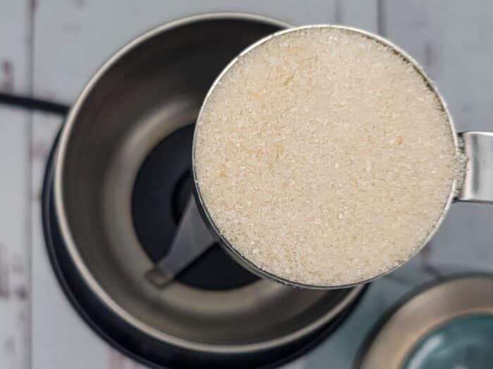 A measuring cup filled with granulated sugar is held above an open food processor.