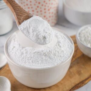 A wooden spoon holding a scoop of white powder over a bowl filled with the same powder, with measuring spoons and additional bowls of powder in the background.