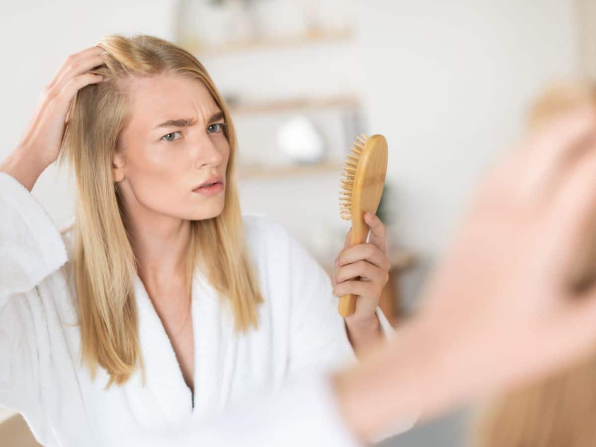 A person with long blonde hair in a white bathrobe looks in a mirror, holding a hairbrush and examining their scalp with a concerned expression.