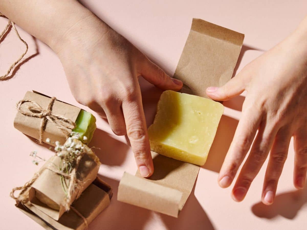 Hands wrapping a bar of yellow soap in brown paper. Two packages of wrapped soap tied with twine and small flowers are beside. The background is light pink.