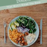 A plate with sliced avocado, corn, spinach, sliced red onions, sausage pieces, and potato salad on a wooden table with a fork and knife.