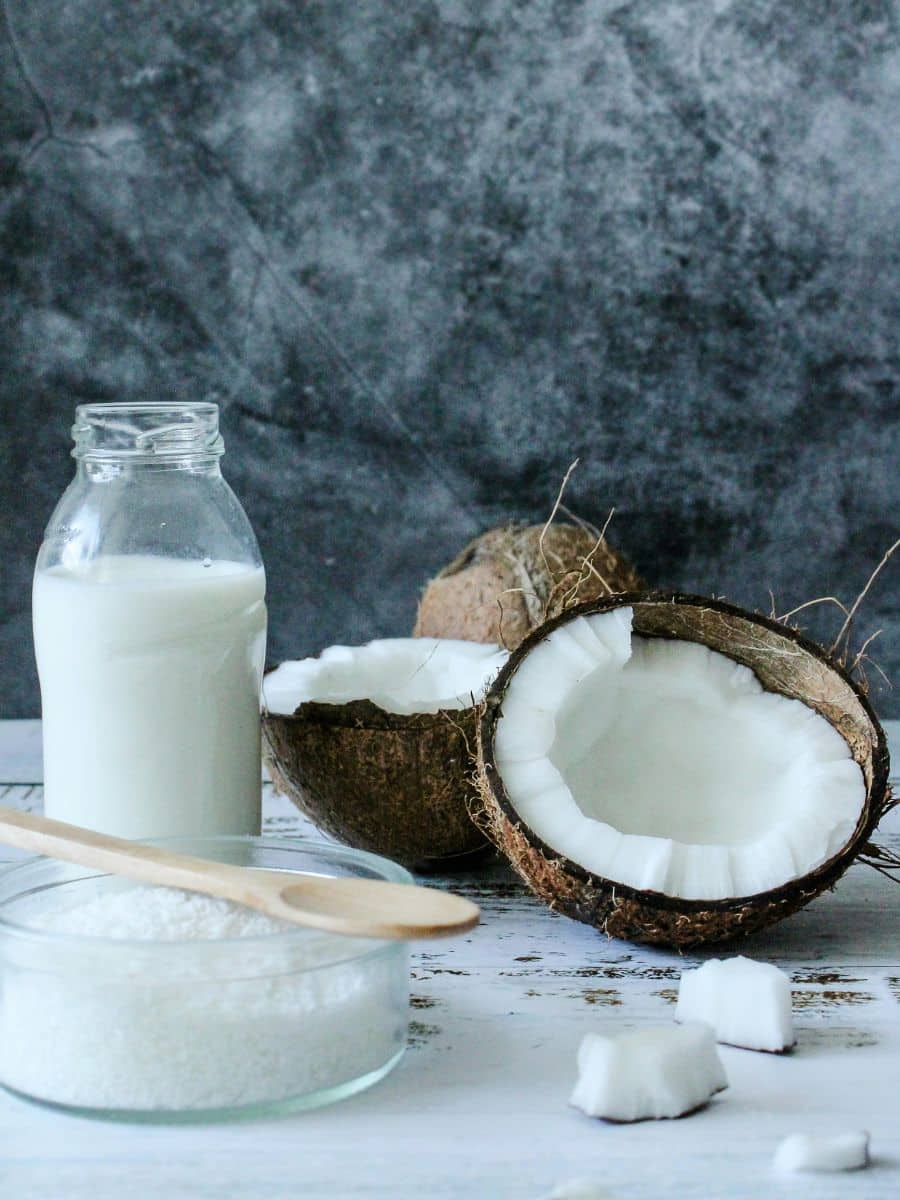 A wooden spoon rests on a bowl of shredded coconut next to halved coconuts and a glass bottle of coconut milk on a white surface, with a textured gray background.