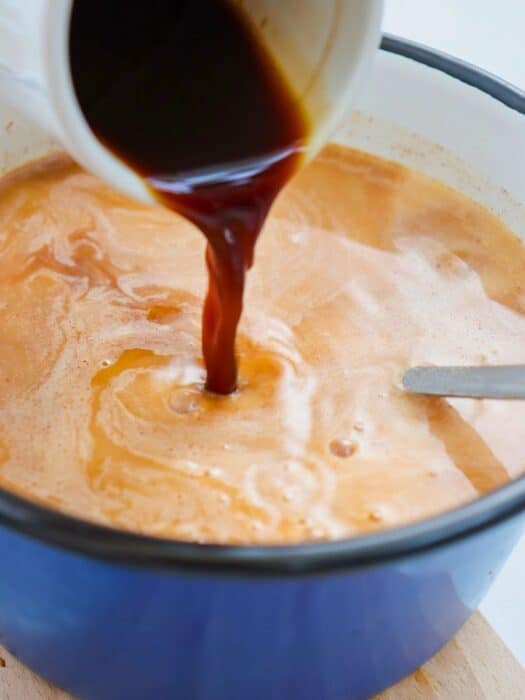 Dark liquid being poured into a pot of light brown mixture, with a spoon partially visible on the right.