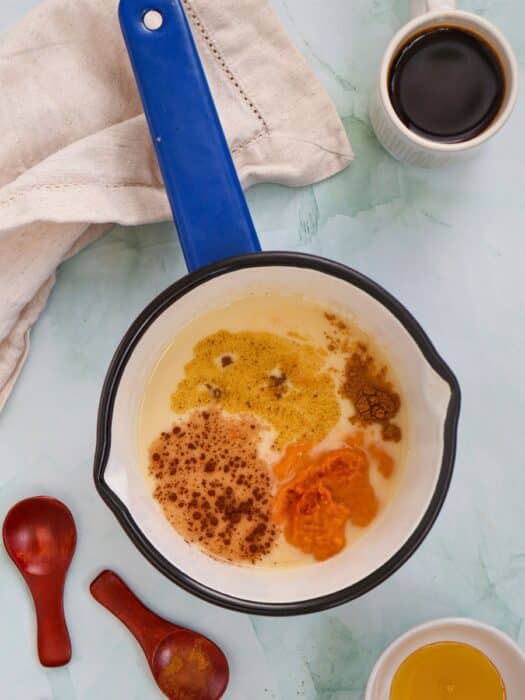 A saucepan with creamy liquid, spices, and pumpkin puree. Nearby are wooden spoons, a beige cloth, a cup of dark liquid, and a small bowl of honey.