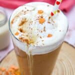 Iced coffee with whipped cream, cinnamon, and heart-shaped sprinkles in a tall glass with a straw on a wooden coaster. Text on image: "How to Make Starbucks-Level PSL at Home.