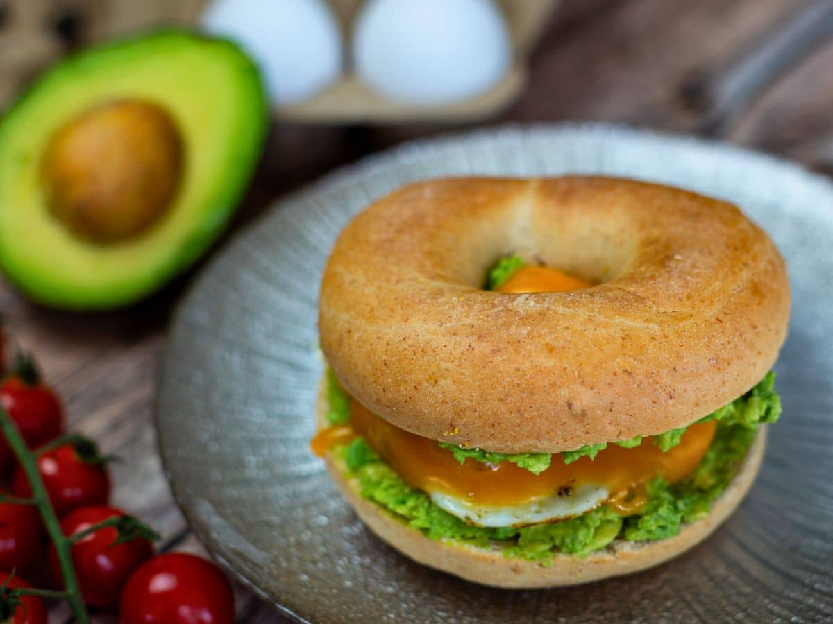 Bagel with avocado, sunny-side-up egg, and cheese on a plate. Avocado half, eggs, and cherry tomatoes in the background.