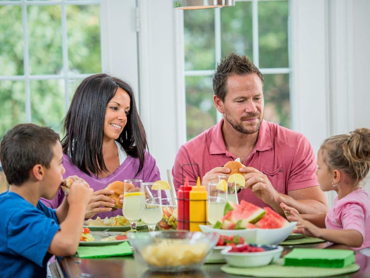 A family of four sits at a table enjoying a meal with burgers, watermelon, and condiments.