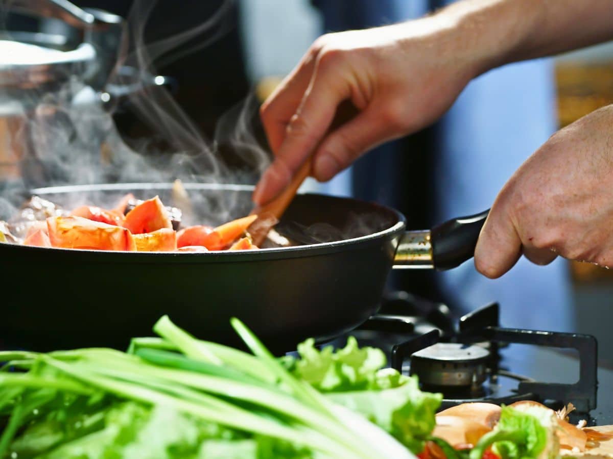 Person cooking vegetables in a frying pan on a stove, with green onions and lettuce nearby.