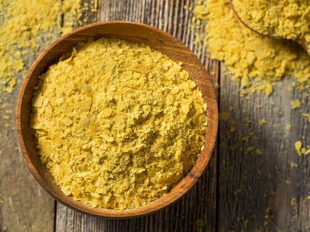 A wooden bowl filled with yellow nutritional yeast flakes on a rustic wooden surface, with some flakes scattered around.