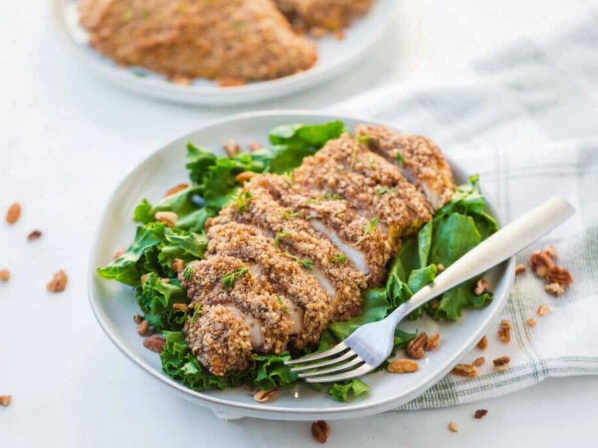 A sliced breaded chicken fillet served on a bed of leafy greens, placed on a white plate with a fork. Some pecans are scattered around.