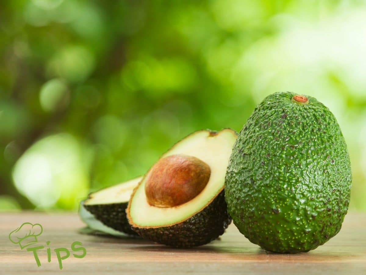 Whole and halved avocados on a wooden surface, with a blurred green background.