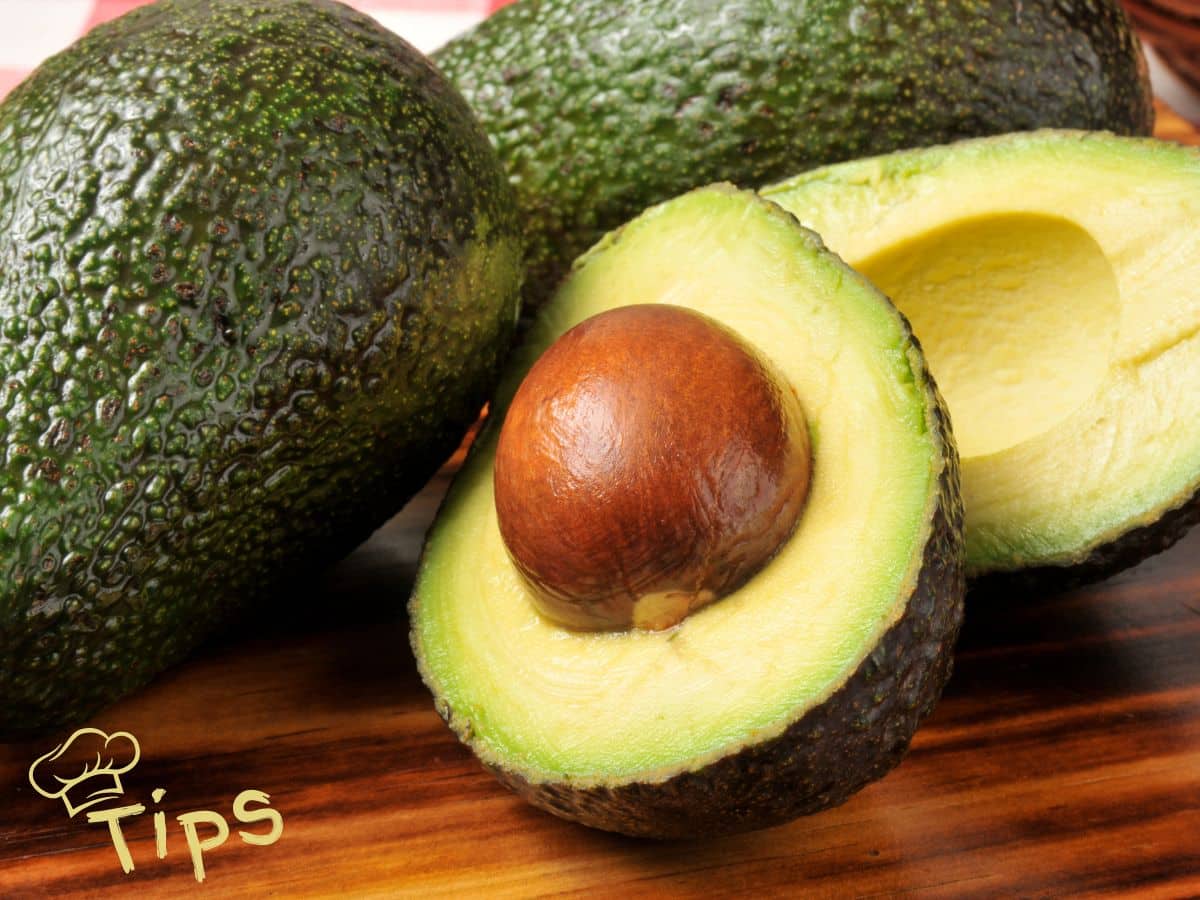 Three avocados on a wooden surface, one of which is cut in half to reveal the seed. "Tips" is written in the corner with a chef's hat graphic.