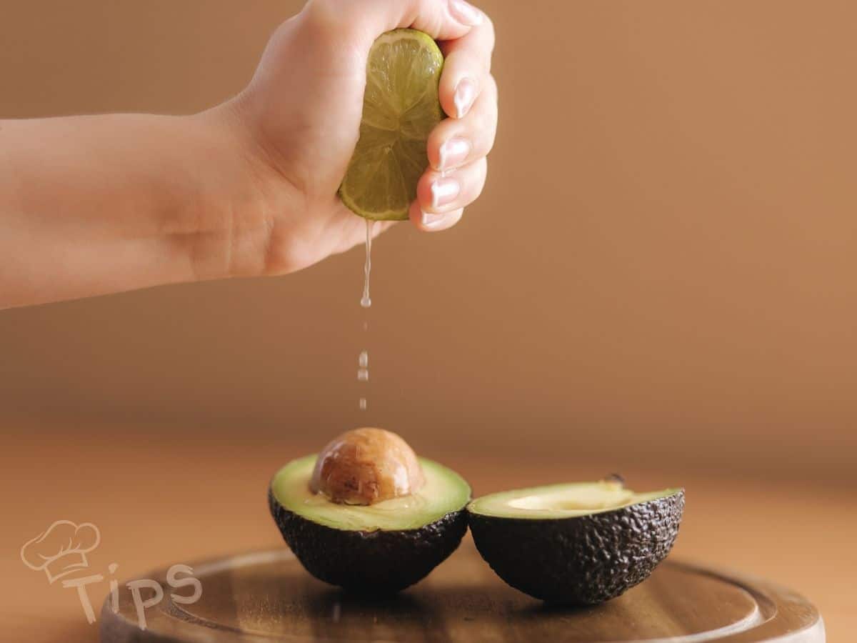 Hand squeezing lime juice over halved avocado on a wooden surface.