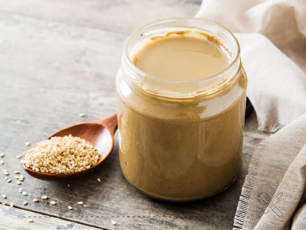 A jar of tahini on a wooden surface, next to a wooden spoon filled with sesame seeds and a beige cloth.