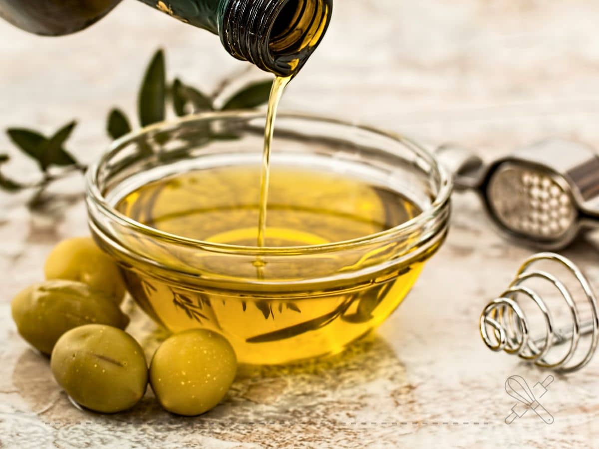 Olive oil being poured into a glass bowl, with green olives and a whisk nearby on a marble surface.