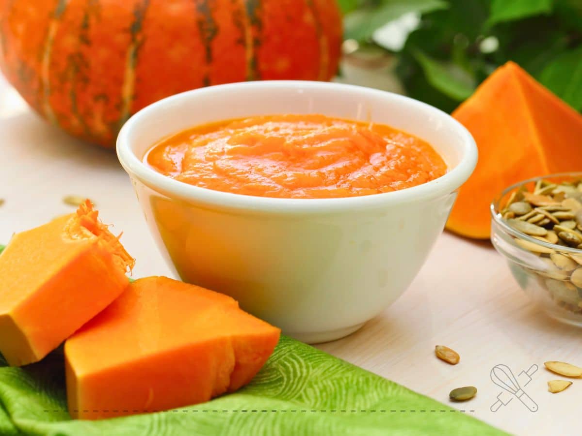 Bowl of orange pumpkin puree surrounded by pumpkin slices, seeds in a small bowl, and a whole pumpkin in the background.