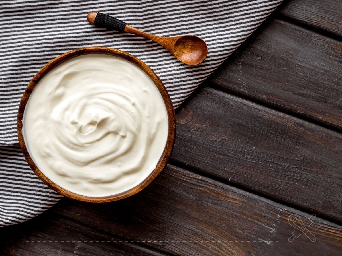 A wooden bowl of yogurt on a dark wooden table, next to a wooden spoon and a striped black and white cloth.