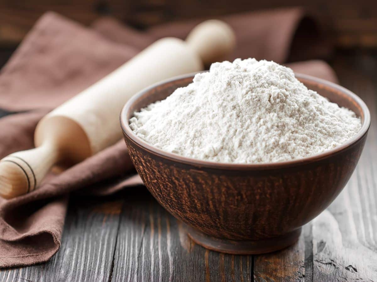 Wooden bowl of flour on wooden table top next to a rolling pin