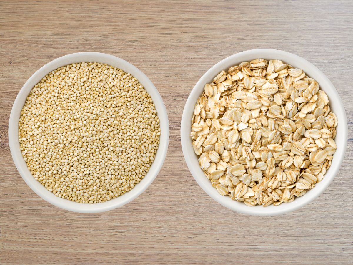 A white bowl of quinoa and a white bowl of oatmeal side-by-side on a wooden table.