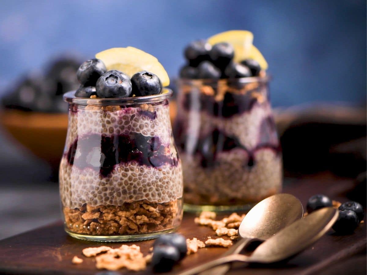 Two glass jars of chia seed pudding layered with blueberries and granola, topped with fresh blueberries and a lemon slice, with two golden spoons beside.