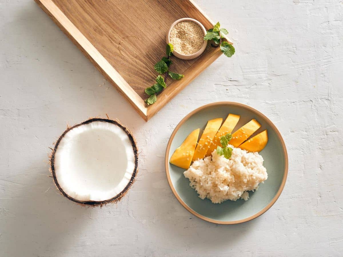 A plate of coconut rice with mango slices next to a halved coconut and a small bowl of sugar on a wooden tray.