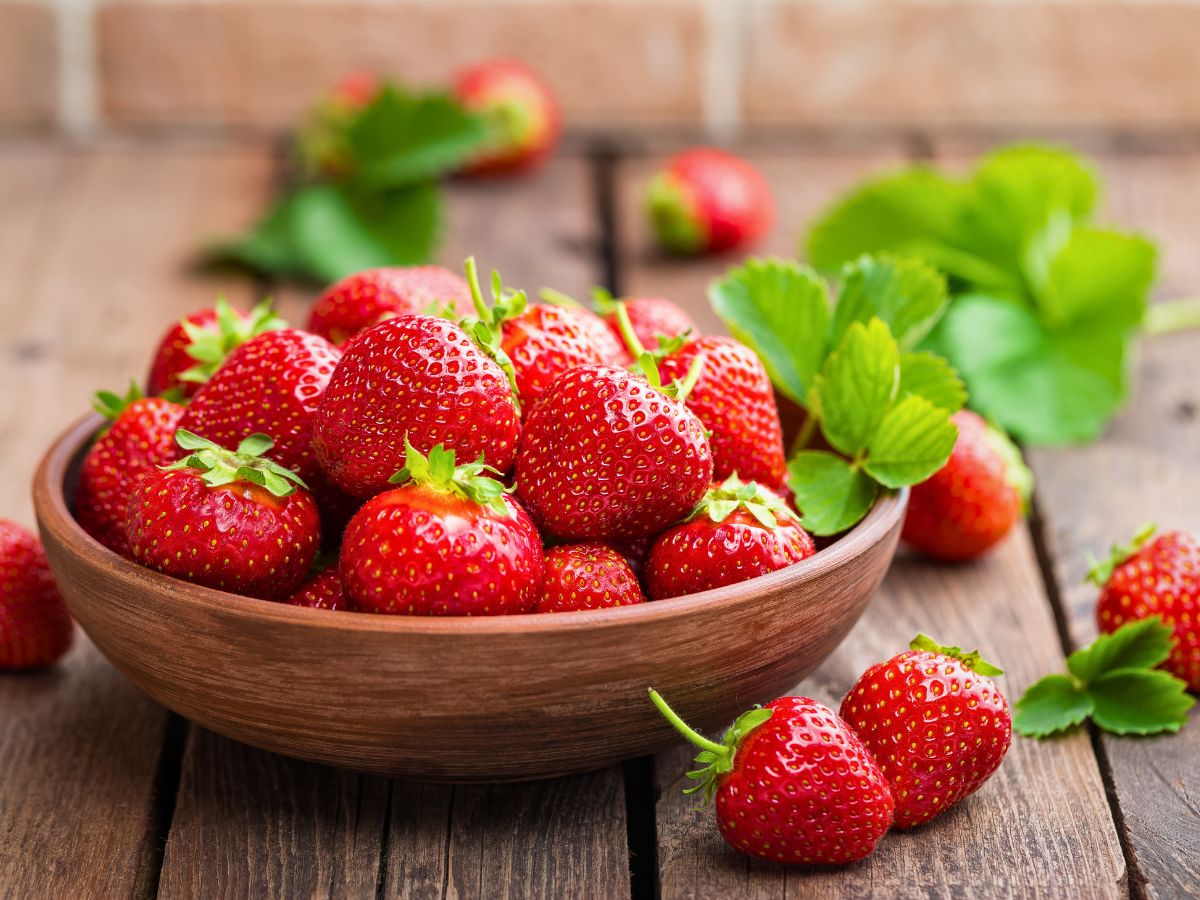 A wooden bowl filled with fresh strawberries sits on a rustic wooden table, with a few strawberries and green leaves scattered nearby.