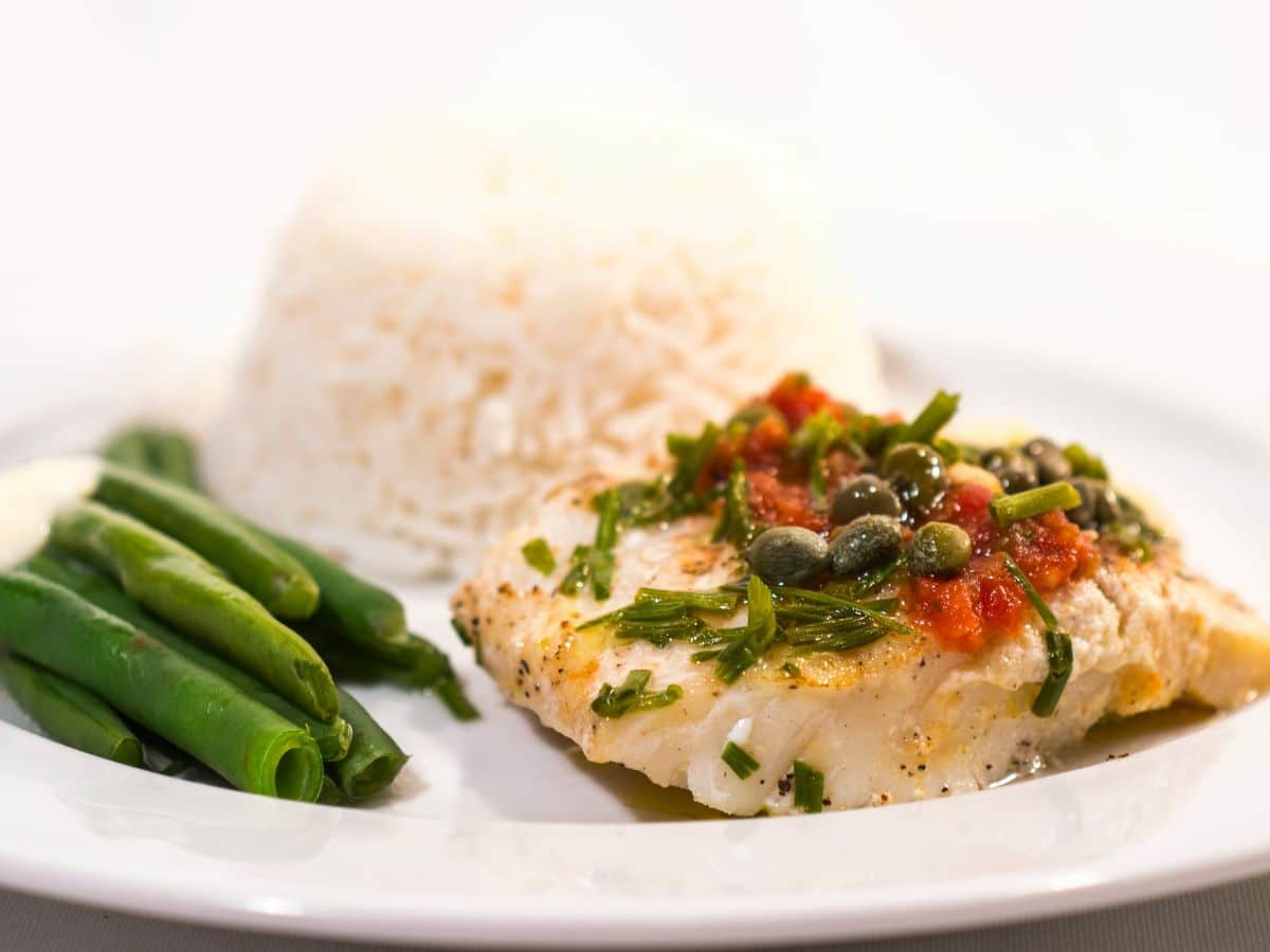 A plate with a portion of white fish topped with capers and herbs, accompanied by green beans and a serving of white rice.