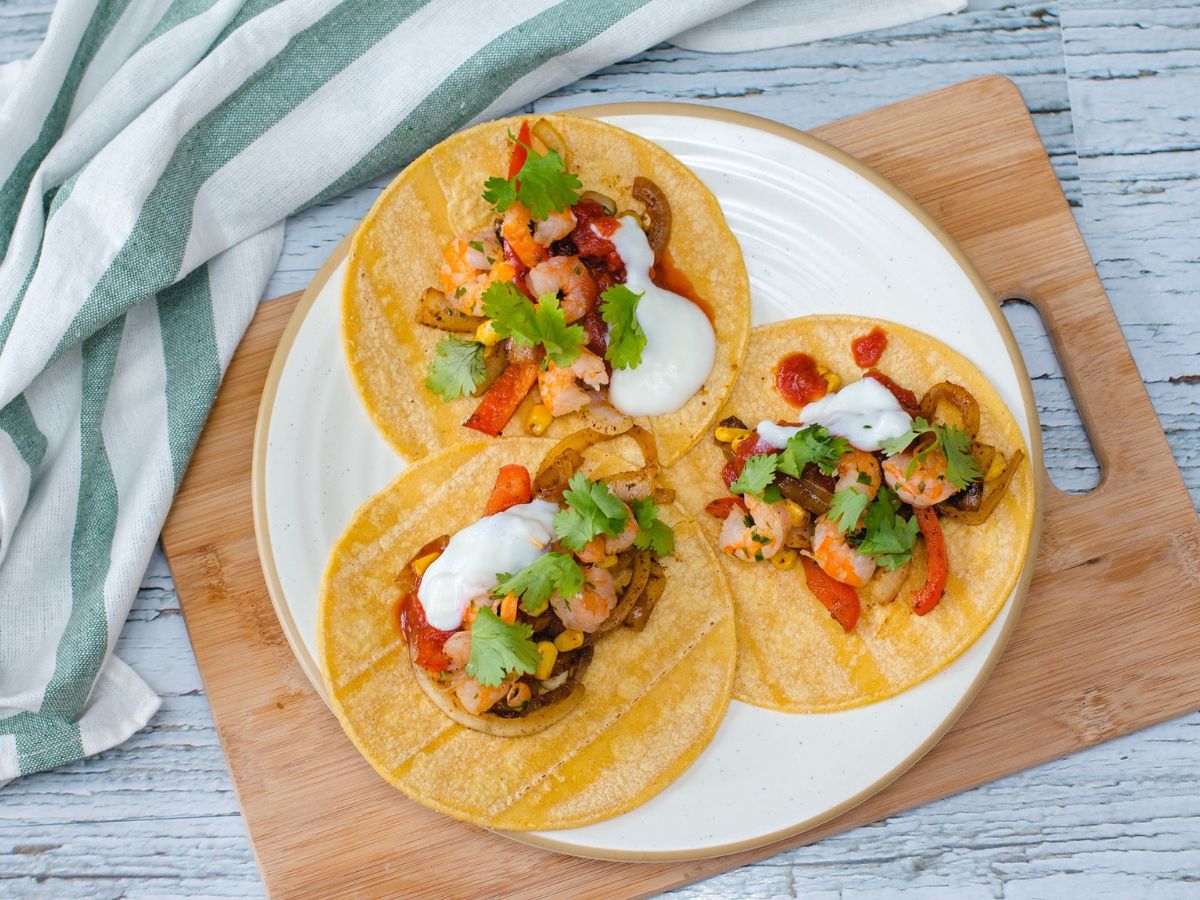 Three shrimp tacos are arranged on a white plate, garnished with cilantro, sour cream, cooked vegetables, and red sauce. The plate is on a wooden cutting board, next to a striped cloth.