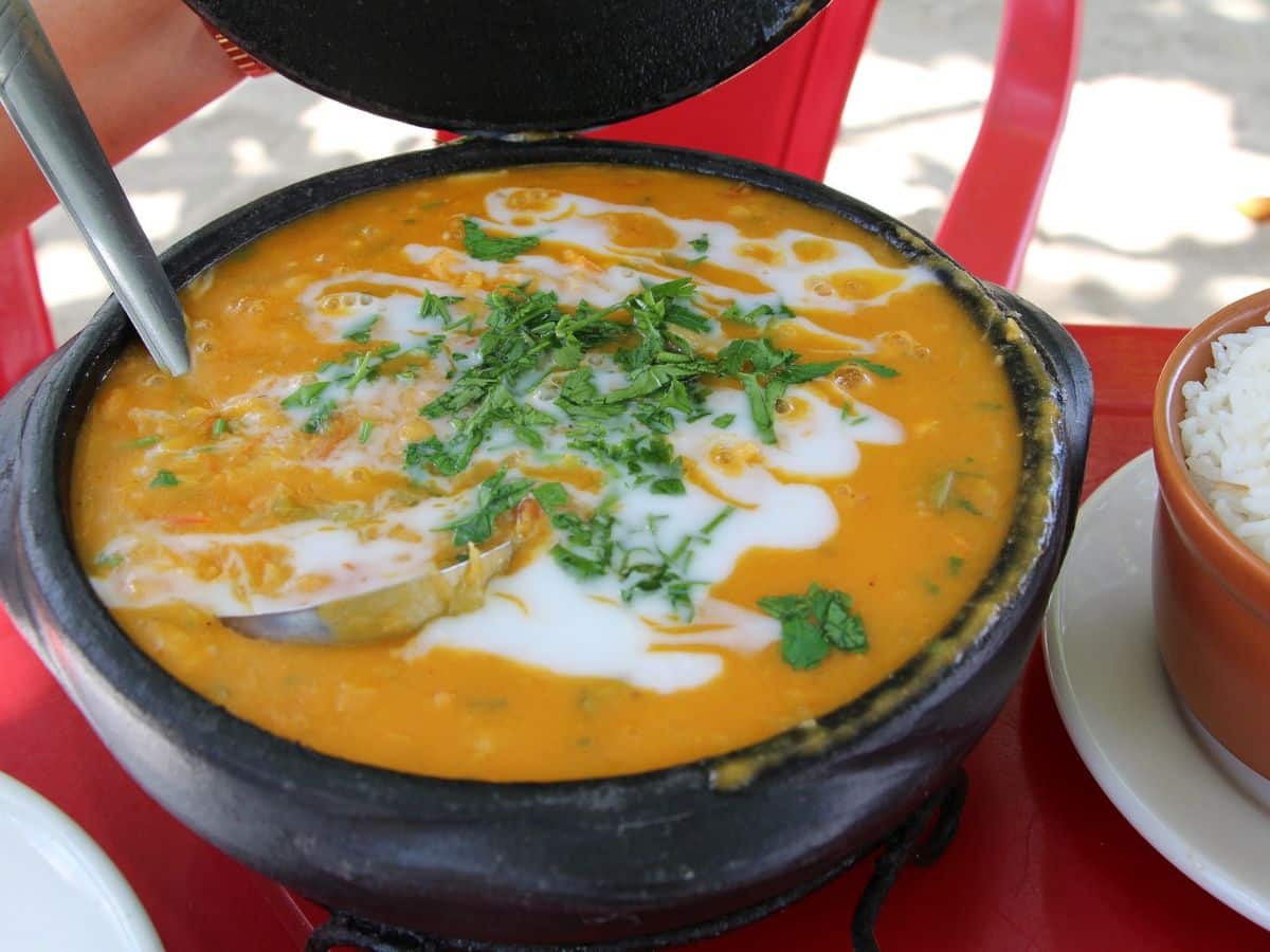 A bowl of orange seafood stew with a garnish of green herbs and white cream, served with a side of white rice in a small bowl on a red table.