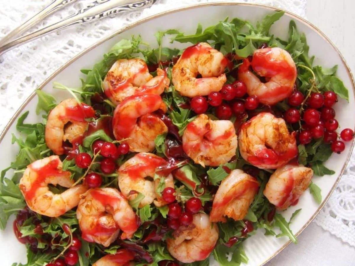 A white oval dish contains shrimp placed on a bed of arugula, garnished with red currants and drizzled with a red sauce, accompanied by a pair of silver forks placed beside the dish.