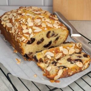 A freshly baked loaf of almond-topped bread with slices revealing chocolate chips, placed on parchment paper on a cooling rack with a knife beside it.