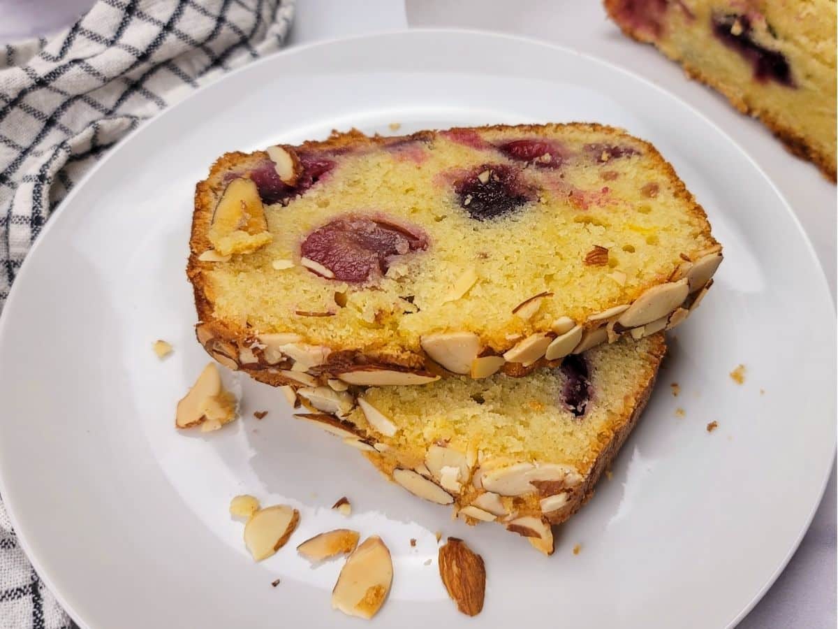 Two slices of cherry loaf cake on a white plate, garnished with sliced almonds, next to a checkered napkin.