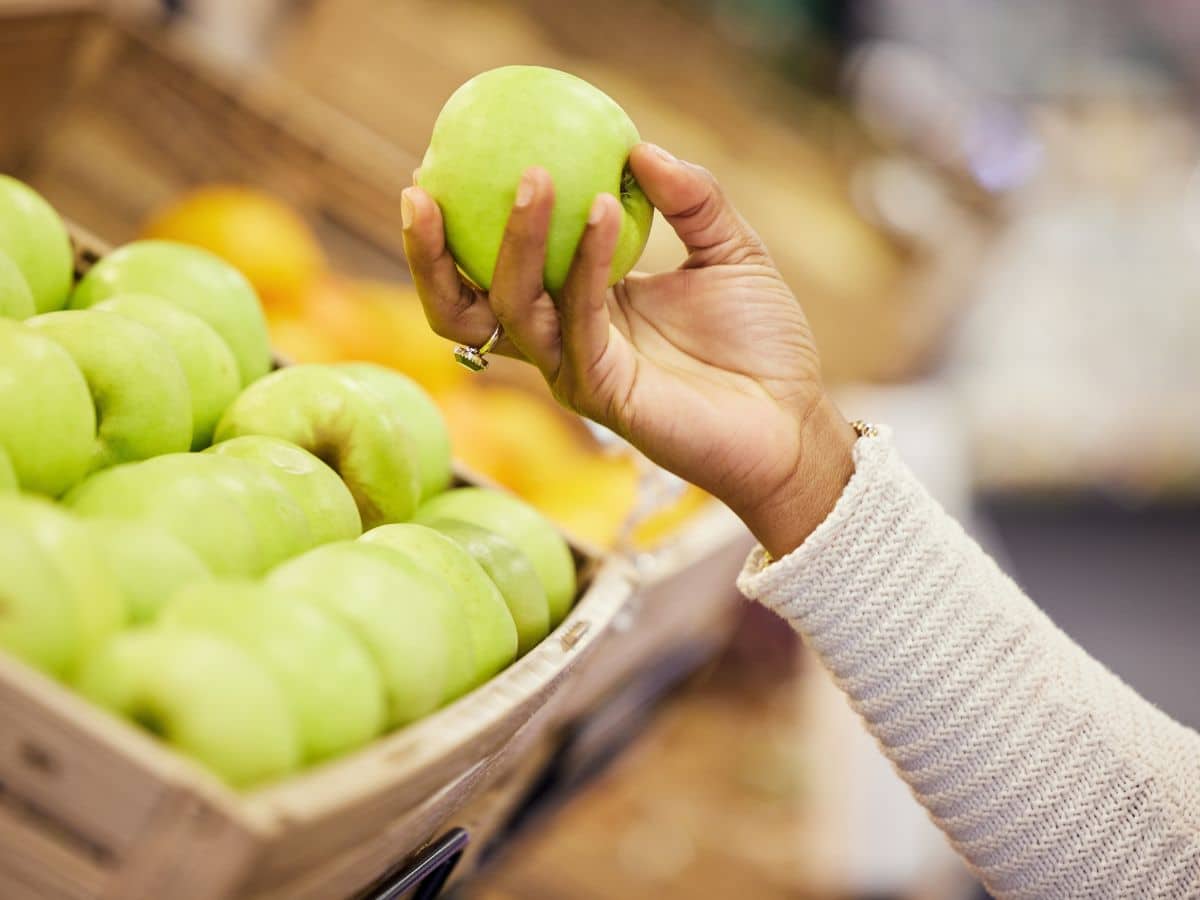 A hand holding a green apple above a display of green apples at a market.