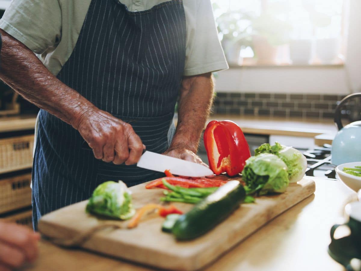 Person wearing an apron chopping vegetables, including bell pepper, zucchini, and lettuce, on a wooden cutting board in a kitchen.