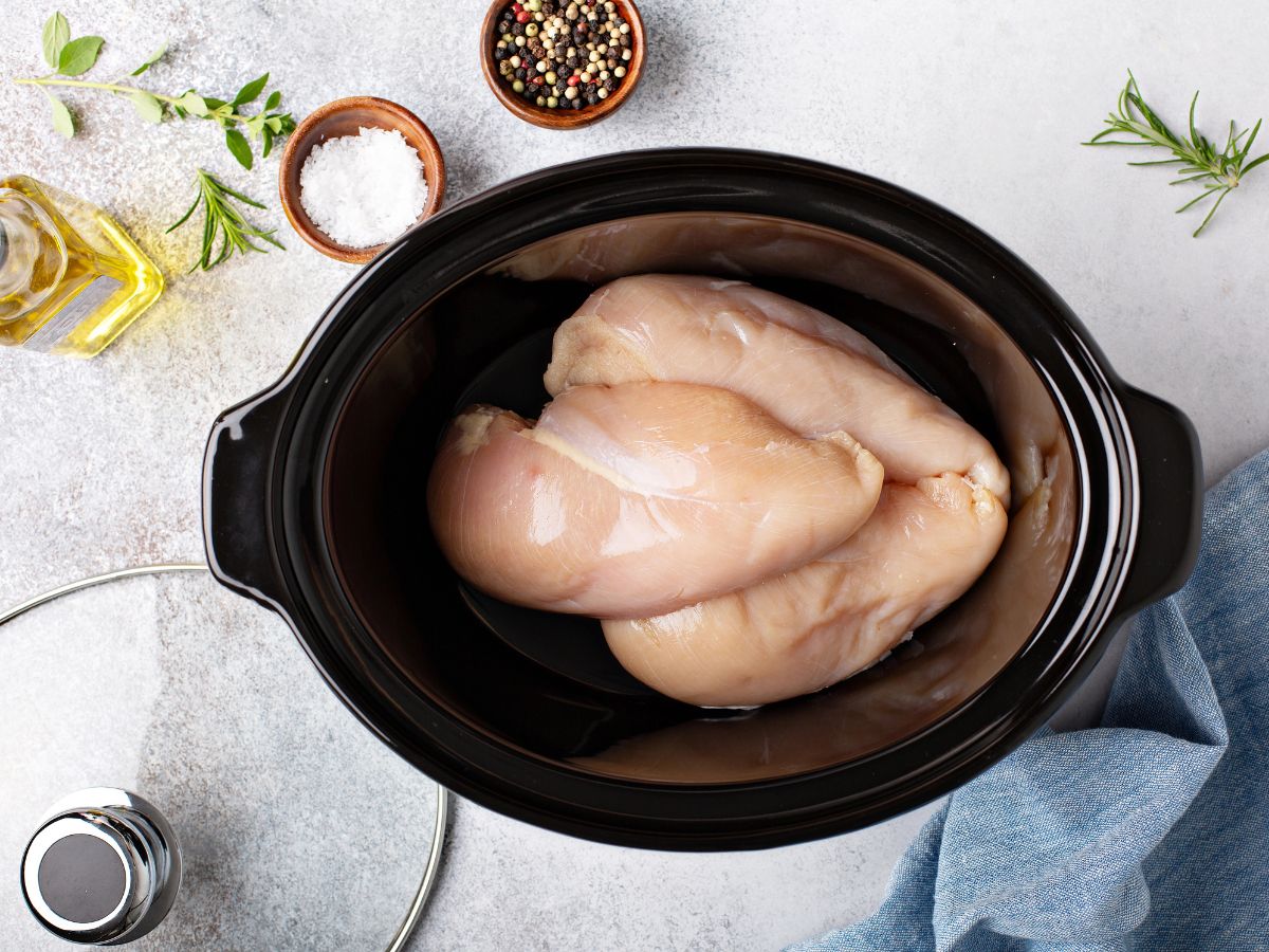Three raw chicken breasts in a slow cooker surrounded by salt, pepper, olive oil, and fresh herbs on a light-colored surface.