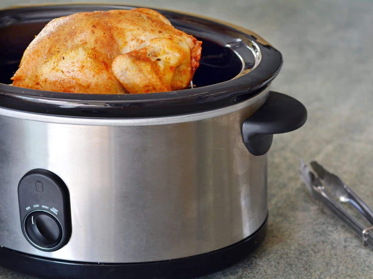 A whole seasoned chicken is cooking in a stainless steel slow cooker. Stainless steel tongs are placed on the countertop next to the slow cooker.