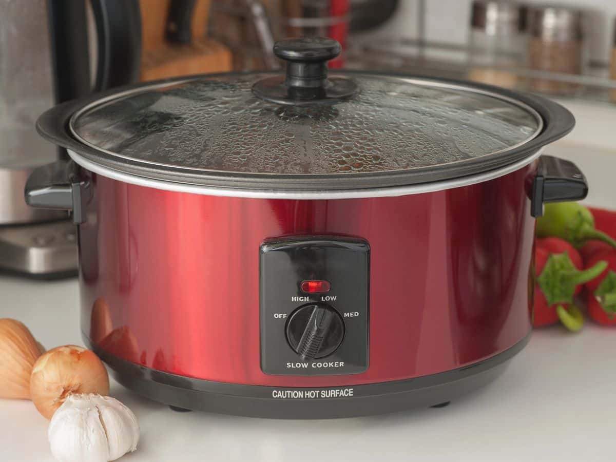 Red slow cooker with a transparent glass lid on a kitchen counter, surrounded by onions, garlic, and bell peppers. The control dial is set to "low.