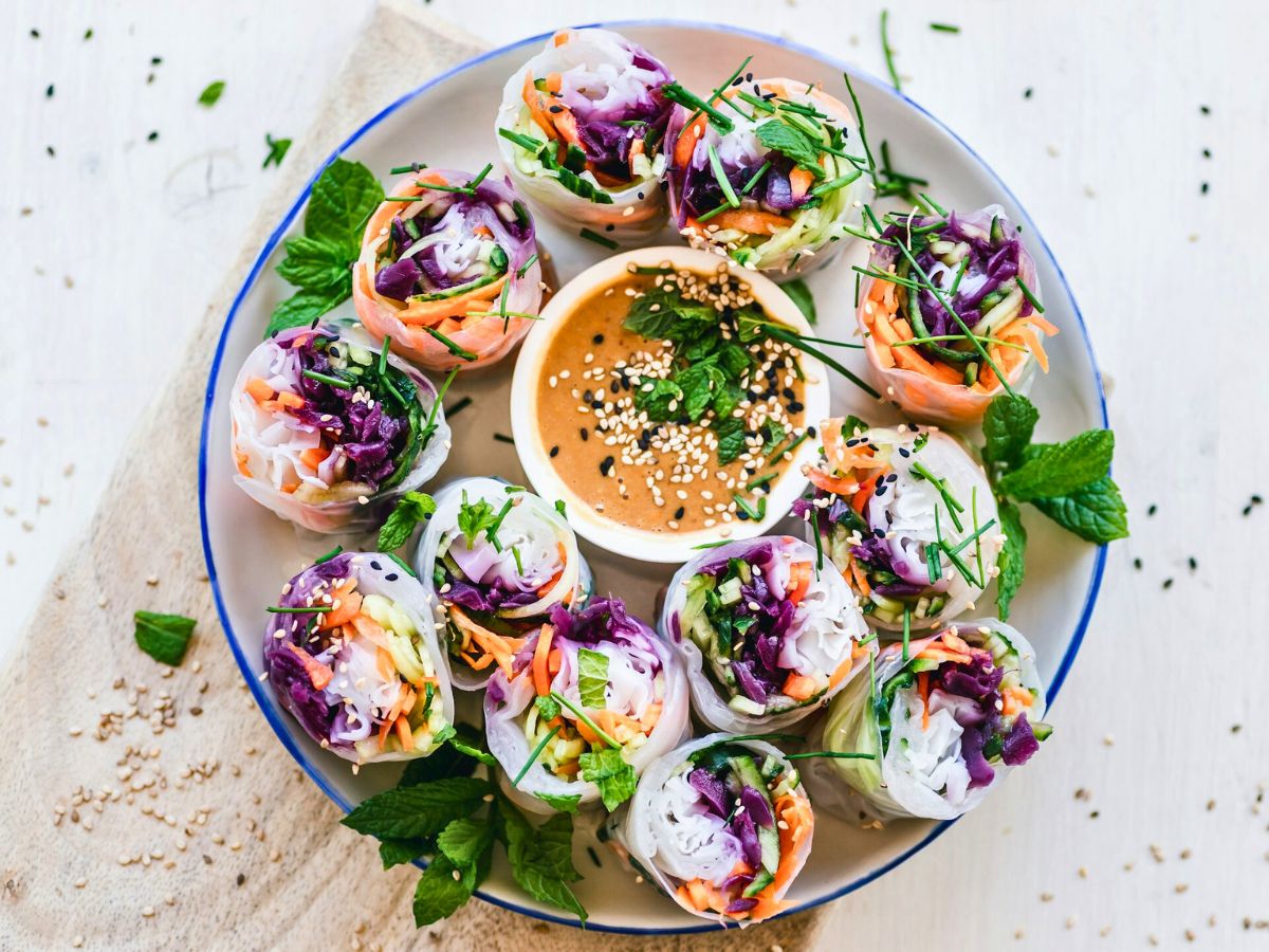 Colorful vegetable spring rolls arranged in a circle on a plate around a bowl of dipping sauce, garnished with fresh herbs and sesame seeds.