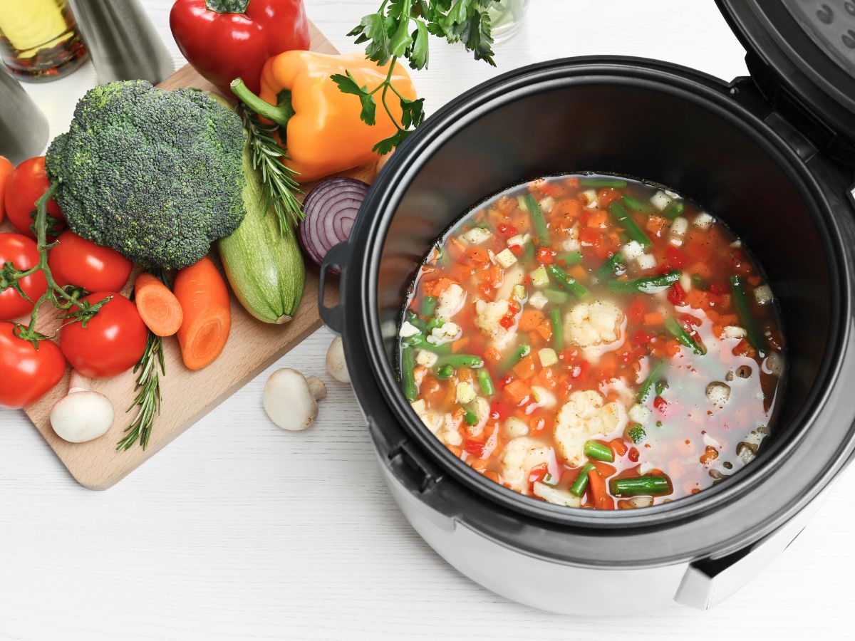 A slow cooker filled with vegetable soup next to a cutting board with fresh vegetables including tomatoes, broccoli, carrots, bell peppers, zucchini, red onion, and mushrooms.
