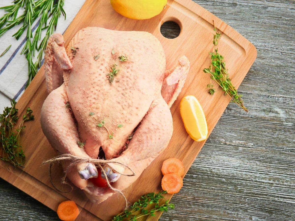 Whole raw chicken with herbs and lemon slices on a wooden cutting board, surrounded by sprigs of rosemary and sliced carrots.
