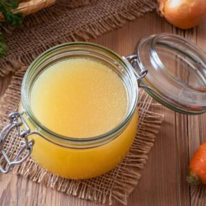 A glass jar of vegetable broth on a wooden table, surrounded by a carrot, an onion, and green herbs.