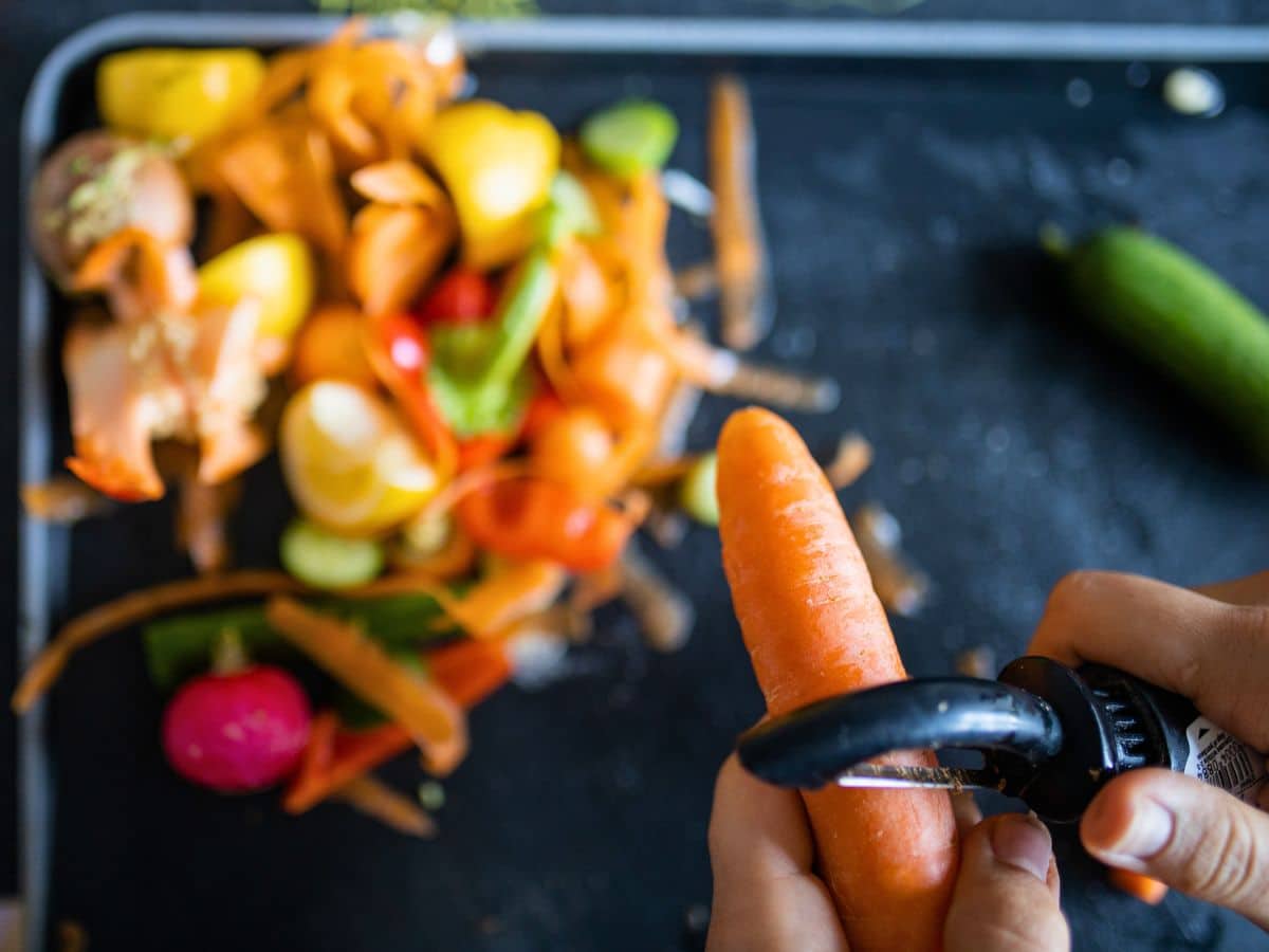 Hands peeling a carrot over a cutting board with assorted vegetable peels and scraps.